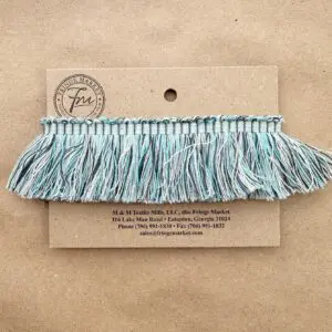 An aqua and white Chatham Cut Fringe tassel decoration attached to a cardboard label from Fringe Market, placed on a textured brown background.